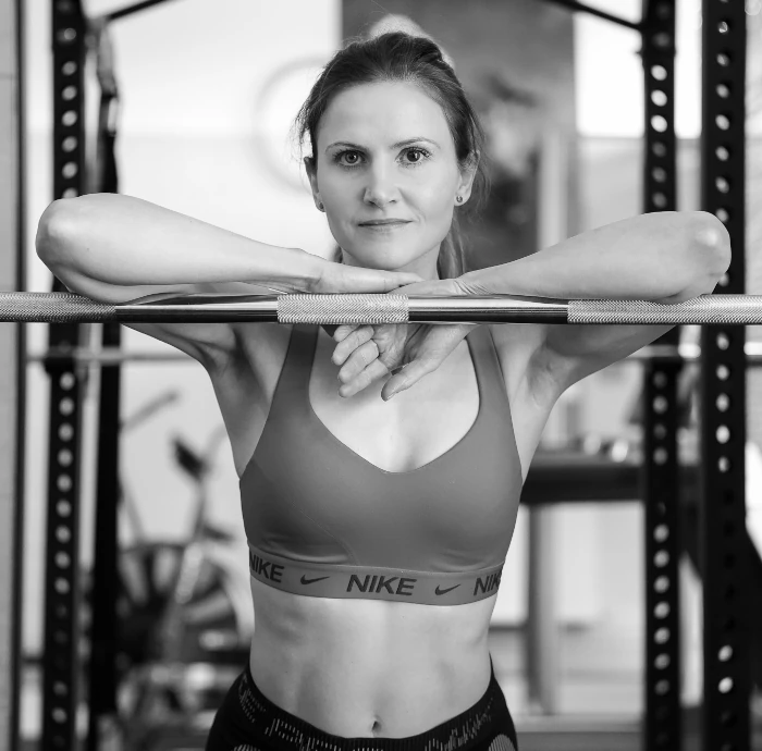 Kobieta opierająca się na sztandze w siłowni, patrząca prosto w obiektyw.